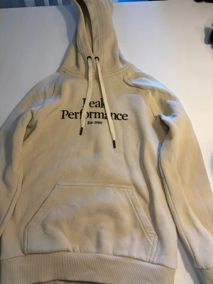 Beige Peak Performance hoodie - Snygg beige hoodie från Peak Performance med stor huva och dragsnören. Klassisk känguruficka framtill och tryckt logga på bröstet. Tillverkad i mjukt bomullsmaterial som känns skönt mot huden. Perfekt för en avslappnad och trendig look.
