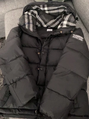 Svart pufferjacka från Burberry - Svart pufferjacka från Burberry med klassiskt rutigt foder i huvan och logga på ärmen. Jackan har dragkedja och knappar framtill samt två fickor med dragkedja. Perfekt för kalla vinterdagar och riktigt snygg streetstil.