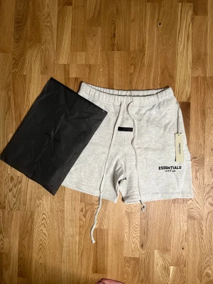 Essentials ljusgrå shorts med snörning - Säljer ett par ljusgrå shorts från Essentials med snörning i midjan och tryckt logga på vänster ben. Shortsen har elastisk midja och är tillverkade i mjuk bomullsmix, perfekta för en avslappnad stil. Levereras med originalpåse och tagg kvar.
