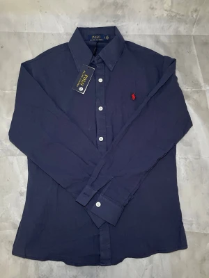 Mörkblå skjorta från Polo Ralph Lauren - Snygg mörkblå skjorta från Polo Ralph Lauren med klassisk krage och långa ärmar. Skjortan har vita knappar och den ikoniska röda logon broderad på bröstet. Perfekt för dig som gillar stilrena plagg med premiumkänsla.