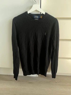 Svart merinoullströja Polo Ralph Lauren - Klassisk svart tröja från Polo Ralph Lauren i slim fit-modell, tillverkad i mjuk och tvättbar merinoull. Tröjan har rund halsringning och den ikoniska broderade loggan på bröstet. Perfekt för en stilren och enkel look.