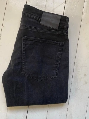 Svarta raka jeans från Jack and Jones - Säljer ett par svarta jeans från Jack and Jones med klassisk femficksmodell och raka ben. Jeansen har en stilren look med svart läderpatch bak i midjan och är tillverkade i ett mjukt denimtyg. Perfekta för dig som gillar minimalistisk stil. Modellen är Tapered Mike. Midjebredden är 40cm. Längden är 94cm. Smalben bredden är 20cm. 