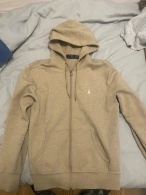 Beige hoodie från Polo Ralph Lauren - Snygg beige hoodie med dragkedja från Polo Ralph Lauren. Klassisk design med huva och dragsko, två fickor framtill och den ikoniska vita loggan broderad på bröstet. Perfekt för en avslappnad och stilren look. Kom med bud i dm