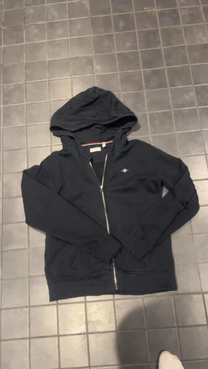 Svart zip-hoodie från Gant - Klassisk svart hoodie med dragkedja från Gant. Tröjan har huva med snörning, två fickor framtill och en liten broderad logga på bröstet. Perfekt för en avslappnad och stilren look.