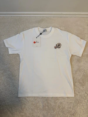 Vit Moncler t-shirt med logga - Snygg vit t-shirt från Moncler med klassisk rund hals och korta ärmar. T-shirten har Moncler-logga på bröstet och en mindre logga på ärmen. Tillverkad i mjuk bomull för en skön känsla och clean look.