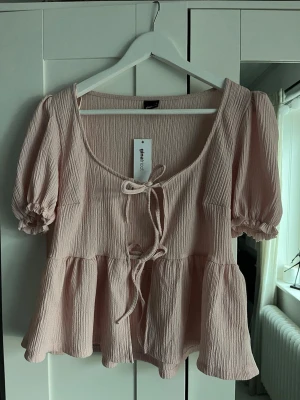 Ljusrosa blus från Gina Tricot - Supersöt ljusrosa blus från Gina Tricot med puffiga korta ärmar och volang nedtill. Blusen har en djup rundad urringning med knytband framtill och är gjord i ett mjukt, krinklat tyg som ger en avslappnad men trendig look. Helt oanvänd med prislapp. Passade inte på mig så säljer. 