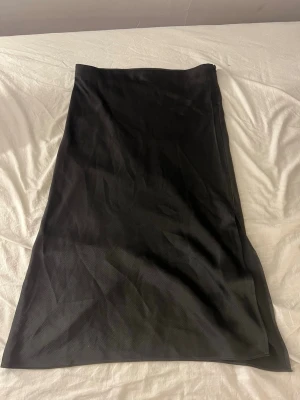 Svart långkjol med dragkedja - En stilren svart långkjol med rak passform och diskret dragkedja i sidan. Kjolen har en slät yta och är perfekt för dig som gillar minimalistisk stil. Passar till många olika looks och är enkel att matcha.