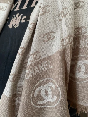 Beige halsduk från Chanell - Snygg beige halsduk från Chanell med ikoniska loggor och text i ljusare och mörkare beige toner. Halsduken har fransade kanter och är tillverkad i ett mjukt, vävt material som känns lyxigt och stilrent. Perfekt accessoar för att lyfta din outfit.