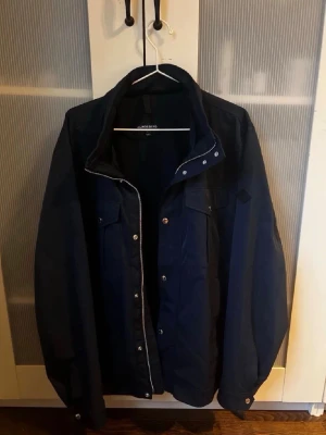 Mörkblå vindjacka från Lindbergh - Stilren svart vindjacka från Lindbergh med dragkedja och tryckknappar framtill. Jackan har två bröstfickor med lock och två sidofickor. Perfekt för blåsiga dagar och enkel att matcha med olika outfits.