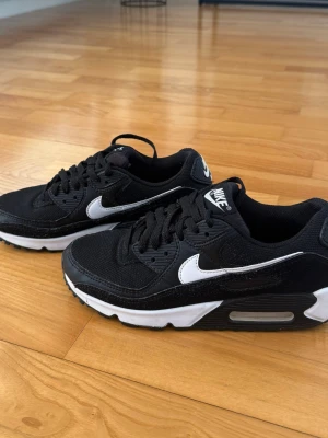 Nike Air Max 90 svarta sneakers - Snygga svarta Nike Air Max 90 sneakers med vit sula och vit swoosh på sidan. Skorna har en klassisk låg siluett, mesh- och mockadetaljer samt synlig Air-enhet i sulan för extra komfort. Perfekta för dig som gillar streetwear och sportig stil. Sparsamt använda.