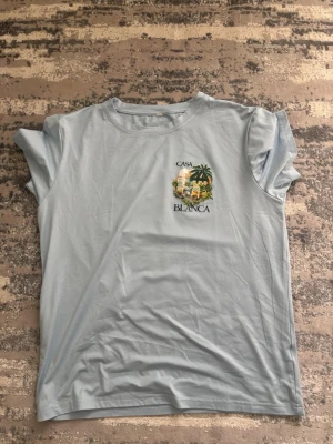 Ljusblå Casa Blanca t-shirt - Snygg ljusblå t-shirt från Casa Blanca med tryck av palmer och byggnader både fram och bak. T-shirten har rund hals och korta ärmar. Perfekt för dig som gillar streetwear och vill sticka ut med ett coolt motiv.