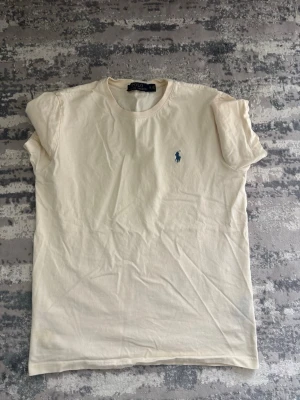 Beige t-shirt från Polo Ralph Lauren - Klassisk beige  t-shirt från Polo Ralph Lauren med rund halsringning och den ikoniska blå logotypen broderad på bröstet. Tillverkad i mjuk bomull som känns skön mot huden. Enkel och stilren design som passar till det mesta.