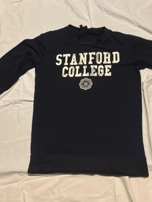 Mörkblå Stanford College sweatshirt - Mörkblå sweatshirt med vit text 'Stanford College' tryckt på bröstet och ett emblem under. Tröjan har rund halsringning och långa ärmar. Tillverkad i mjukt bomullsliknande material, perfekt för en avslappnad och sportig stil.