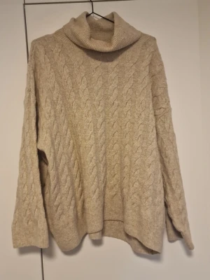 Beige kabelstickad polotröja - Mysig beige kabelstickad polotröja med bred krage och långa ärmar. Tröjan har ett klassiskt flätmönster och ribbade muddar vid ärmslut och nederkant. Perfekt för kalla dagar och enkel att matcha med jeans eller kjol.