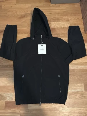Svart vindjacka med huva från Moncler - Svart vindjacka från Moncler med huva och hel dragkedja framtill. Jackan har två fickor med dragkedja och en Moncler-logga på ärmen. Tillverkad i lätt syntetmaterial, perfekt för blåsiga dagar. Enkel och stilren design som passar till det mesta. Storlek är S och dragkedjan där nere är lite sönder men går att få igen med lite arbete! 
