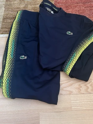 Svart Lacoste set med gula detaljer S-m - Snyggt svart set från Lacoste med matchande tröja och byxor. Båda plaggen har gula och gröna mönstrade ränder längs sidorna och den klassiska Lacoste-loggan på bröstet och benet. Perfekt för en sportig och trendig look.