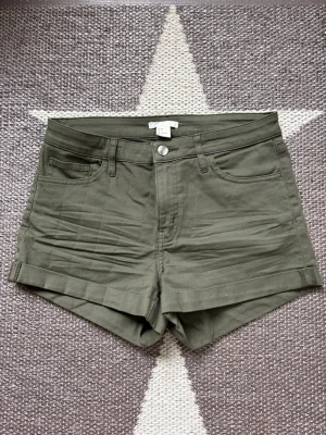 Olivgröna shorts från H&M - Snygga olivgröna shorts från H&M med klassisk femficksdesign och uppvikta benslut. De har knapp- och dragkedjestängning samt bälteshällor. Storlek S