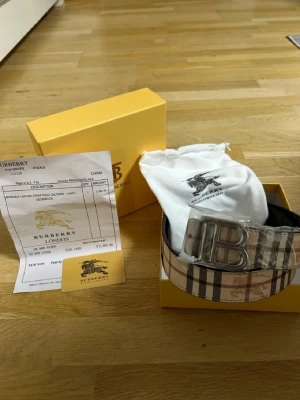  bälte med klassiskt rutmönster - Snyggt  bälte med det ikoniska rutiga mönstret i beige, svart och rött. Bältet har ett stort silvrigt spänne med  logotyp och detaljer i skinn. Perfekt accessoar för att lyfta din outfit.