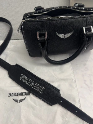 Zadig & Voltaire svart handväska - Cool svart handväska i grained läder från Zadig & Voltaire med silverfärgade metalldetaljer, vingar framtill och bred axelrem med broderad VOLTAIRE-text. Väskan har dubbla handtag och dragkedja upptill.