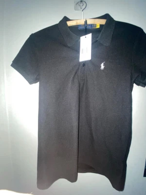 Svart pikétopp från Ralph Lauren - Klassisk svart pikétopp från Ralph Lauren med korta ärmar och vit broderad logga på bröstet. Tidlös design med krage och knappslå framtill. Perfekt till jeans eller kjol för en clean och sportig look.Den passar både Xs och S.Och priset kan diskuteras