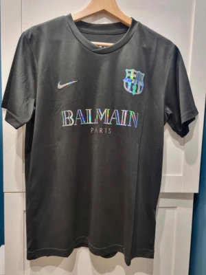 Barcelona Balmain T-Shirr - Snygg svart Barcelona bortatröja från Nike med grått mönster och svart. Tröjan har klubbmärke och Nike -logga på bröstet. Tillverkad i lätt och ventilerande material, perfekt för fotbollsträning eller match.