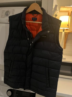 Svart dunväst från Gant - Snygg svart dunväst från Gant med orange foder. Västen har ståkrage, dragkedja och tryckknappar framtill samt två fickor med dragkedja. Perfekt för lager på lager och ger en sportig look.