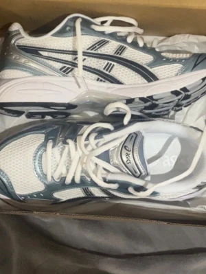 Vita och silver Asics sneakers - Snygga Asics sneakers i vitt och silver med klassiska svarta detaljer och mesh-ovandel för bra andningsförmåga. Skorna har en sportig vibe med tydliga Asics-loggor på sidorna och en bekväm, vadderad sula. Perfekta för dig som gillar streetstyle och sportig look.