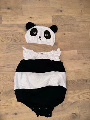 Panda maskeraddräkt med mössa - Söt panda maskeraddräkt med svartvita ränder och tillhörande mössa med öron och ansikte. Dräkten har puffiga ärmar och knappar i grenen. Perfekt för maskerad eller cosplay. Materialet känns mjukt och bekvämt.