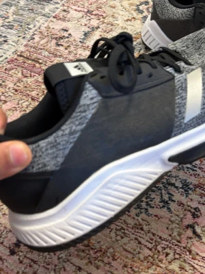 Svarta och grå sneakers från Adidas - Snygga sneakers från Adidas i svart och grått med vita detaljer och vit sula. Skorna har klassisk snörning och en sportig design med diskret Adidas-logga på hälen. Ovandelen är i textil och syntetmaterial för en modern look.