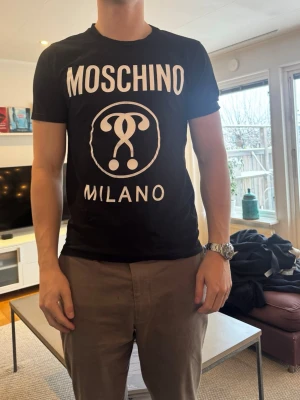Svart Moschino Milano t-shirt - Snygg svart t-shirt från Moschino med stort vitt tryck framtill där det står 'Moschino Milano' och en ikonisk logga. Klassisk passform och rund hals. Perfekt för dig som vill ha en statement tee från ett trendigt märke. 