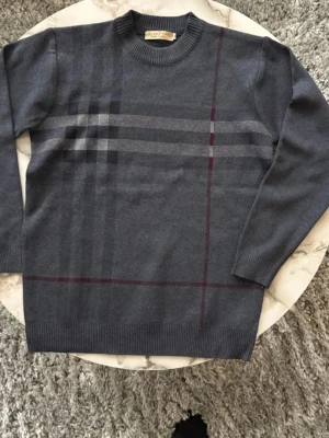 Burberry stickad tröja - Snygg grå stickad tröja från Burberry med klassiskt rutmönster i mörkgrått och vinrött. Rund halsringning och långa ärmar med ribbade muddar. Perfekt för dig som gillar stilrena och tidlösa plagg med en touch av lyx. Otroligt skön att ha på sig och väldigt bra passform, helt ärligt 10/10 sweatshirt