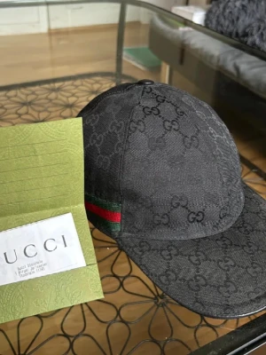Svart keps från Gucci med GG-mönster - Snygg svart keps från Gucci med klassiskt GG-monogram över hela kepsen. Kepsen har en böjd skärm och en grön-röd rand på sidan som ger en exklusiv touch. Tillverkad i canvasmaterial som känns både lyxigt och slitstarkt.