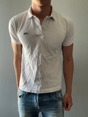 Ljusrosa pikétröja från Lacoste - Klassisk vit pikétröja från Lacoste med korta ärmar och den ikoniska gröna krokodilloggan på bröstet. Tröjan har krage och knäppning med två knappar. Perfekt till jeans eller shorts för en clean och sportig look.