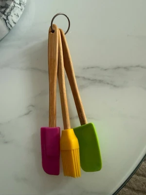 Slickepottar och pensel i silikon - Tre köksredskap med trähandtag och färgglada silikonhuvuden: en rosa slickepott, en gul pensel och en grön slickepott. Alla sitter ihop med en metallring, perfekta för bakning och matlagning.