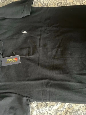 Svart pikétröja från Polo Ralph Lauren - Klassisk svart pikétröja från Polo Ralph Lauren med vit broderad logga på bröstet. Tröjan har krage, knappar framtill och är tillverkad i mjuk bomull. Perfekt för en stilren och avslappnad look.