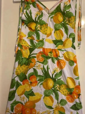 Somrig klänning med fruktmönster - Färgglad klänning med tropiskt fruktmönster i gult, orange och grönt. Klänningen har smala axelband, v-ringning och är ärmlös. Perfekt för varma dagar och ger en fräsch, livlig look.