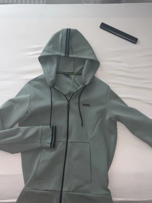 Grått loungewear-set från BOSS - Snyggt och bekvämt loungewear-set från BOSS i ljusgrå färg. Setet består av en hoodie med dragkedja, svarta detaljer och matchande byxor med dragsko i midjan. Båda plaggen har BOSS-logga framtill. Perfekt för chill dagar hemma.