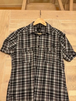 Svartvitrutig skjorta Jack & Jones XL - Kortärmad skjorta från Jack & Jones i svartvitt rutigt mönster. Skjortan har två bröstfickor med knappar och klassisk krage. Perfekt för en avslappnad stil och lätt att matcha med jeans eller shorts.