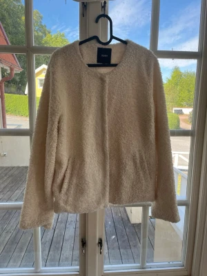 Krämvit teddyjacka från Object - Mysig krämvit teddyjacka från Object med rund halsringning och två sidofickor. Jackan har en fluffig och mjuk yta i teddy-material som ger en avslappnad och trendig look. Perfekt för kyliga dagar när du vill vara både varm och snygg.
