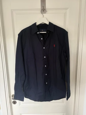Mörkblå skjorta från Ralph Lauren - Klassisk mörkblå skjorta från Ralph Lauren med vit knappstängning och röd broderad logga på bröstet. Skjortan har lång ärm och normal passform, perfekt för en stilren look. Tillverkad i mjuk bomull för skön känsla.