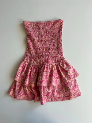 Rosa volangklänning från Gina Tricot - Supersöt kort klänning från Gina Tricot i rosa med blommigt mönster med volangkjol. Klänningen är axelbandslös med smock upptill och har en luftig passform, perfekt för varma dagar. Storlek XS