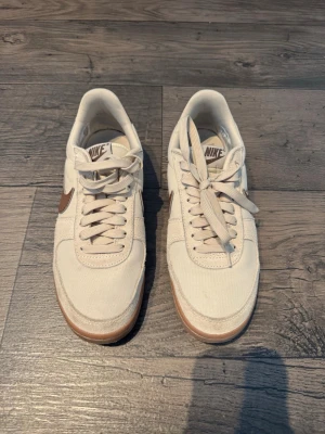 Beige Nike sneakers med bruna detaljer - Snygga beige sneakers från Nike med bruna swoosh-loggor på sidorna och matchande bruna detaljer vid hälen. Skorna har en klassisk låg siluett, vita snören och en gummisula i ljusbrunt. Ovandelen är i textil och mocka för en stilren och trendig look.