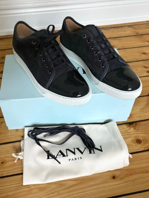 Lanvin svarta sneakers med lackdetalj - Helt nya och oanvända. Snygga svarta sneakers från Lanvin med ovandel i mocka och en glansig lackad tå. Skorna har vita sulor, svarta skosnören och diskreta metalldetaljer vid snörningen. Kommer med originalpåse och extra skosnören.