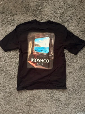 Svart MONACO t-shirt med tryck - Svart t-shirt från Sixth June med texten MONACO på bröstet och ett stort färgglatt tryck på ryggen som föreställer ett fönster med utsikt över havet. Klassisk passform och rund halsringning. Perfekt för dig som gillar streetwear och snygga prints.