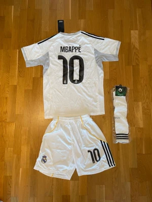Mbappé Real Madrid hemmaställ - Säljer ett komplett Real Madrid matchställ med Mbappé 10-tryck. Setet innehåller vit tröja med svarta detaljer, shorts med klubbmärke och nummer, samt vita strumpor. Materialet är lätt och andas, perfekt för fotbollsträning eller match.