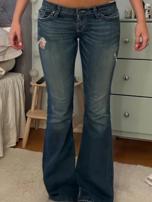 Blå bootcut jeans med slitningar - Köpta för 1400kr. Snygga blå bootcut jeans med slitningar på låret och fransade bakfickor. Jeansen har låg midja och klassisk femficksdesign. Perfekta för dig som gillar en retro vibe och vill sticka ut lite extra.