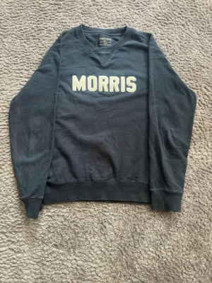 Morris mörkblå sweatshirt - Mörkblå sweatshirt från Morris med stort vitt broderat logotyptryck på bröstet. Tröjan har rund halsringning, ribbade muddar vid ärmslut och nederkant samt klassisk college-look. Perfekt för dig som gillar stilrena och sportiga plagg. Är en storlek L men skulle säga att den passar bättre som en M 