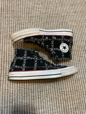 Converse x JW Anderson svarta highs - Snygga svarta Converse x JW Anderson sneakers med vitt och rött mönster, högt skaft och klassisk vit gummisula. Skorna har JW Anderson-tryck över hela ovandelen och Converse All Star-logga på sidan. Perfekta för dig som vill sticka ut med en unik collab.