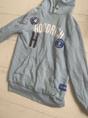 Ljusblå hoodie från Hoodrich XS - Snygg ljusblå hoodie från Hoodrich med vita och svarta tryck och patches på bröstet. Stor magficka framtill och huva med dragsko. Mjuk insida och coola detaljer som broderade märken och logga på ärmen. Perfekt för en avslappnad streetstyle.