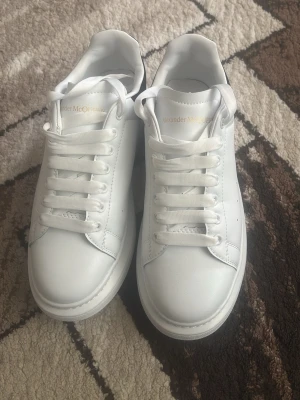 Vita Alexander McQueen sneakers - Stilrena vita sneakers från Alexander McQueen med klassisk låg siluett. Skorna har tjock sula med unikt mönster undertill och är tillverkade i slätt läder. Guldtext på plösen och breda vita skosnören ger en lyxig känsla.